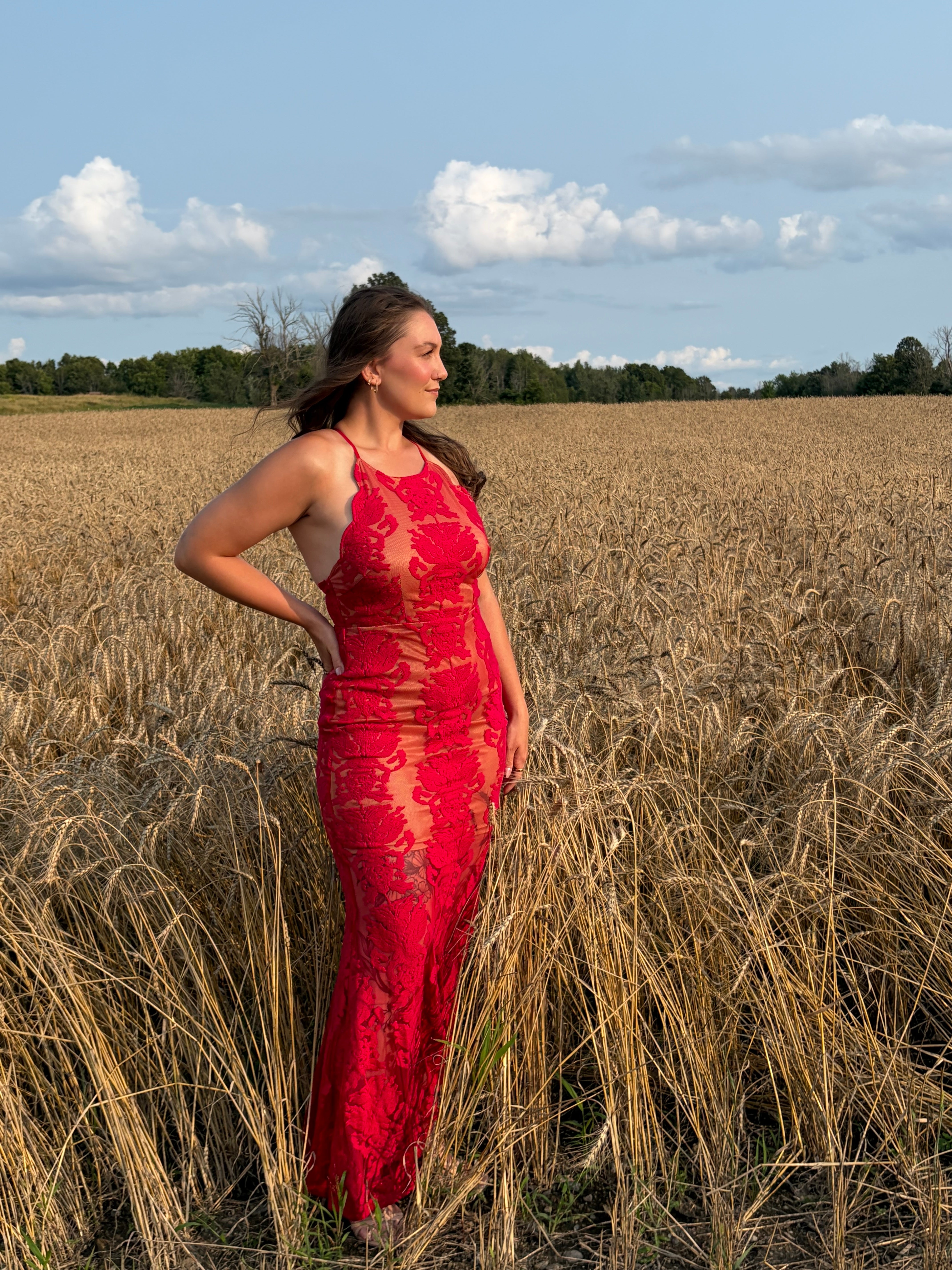 A woman wearing a red lace halter maxi dress stands in a field of dry golden grass under a blue sky with white clouds.
